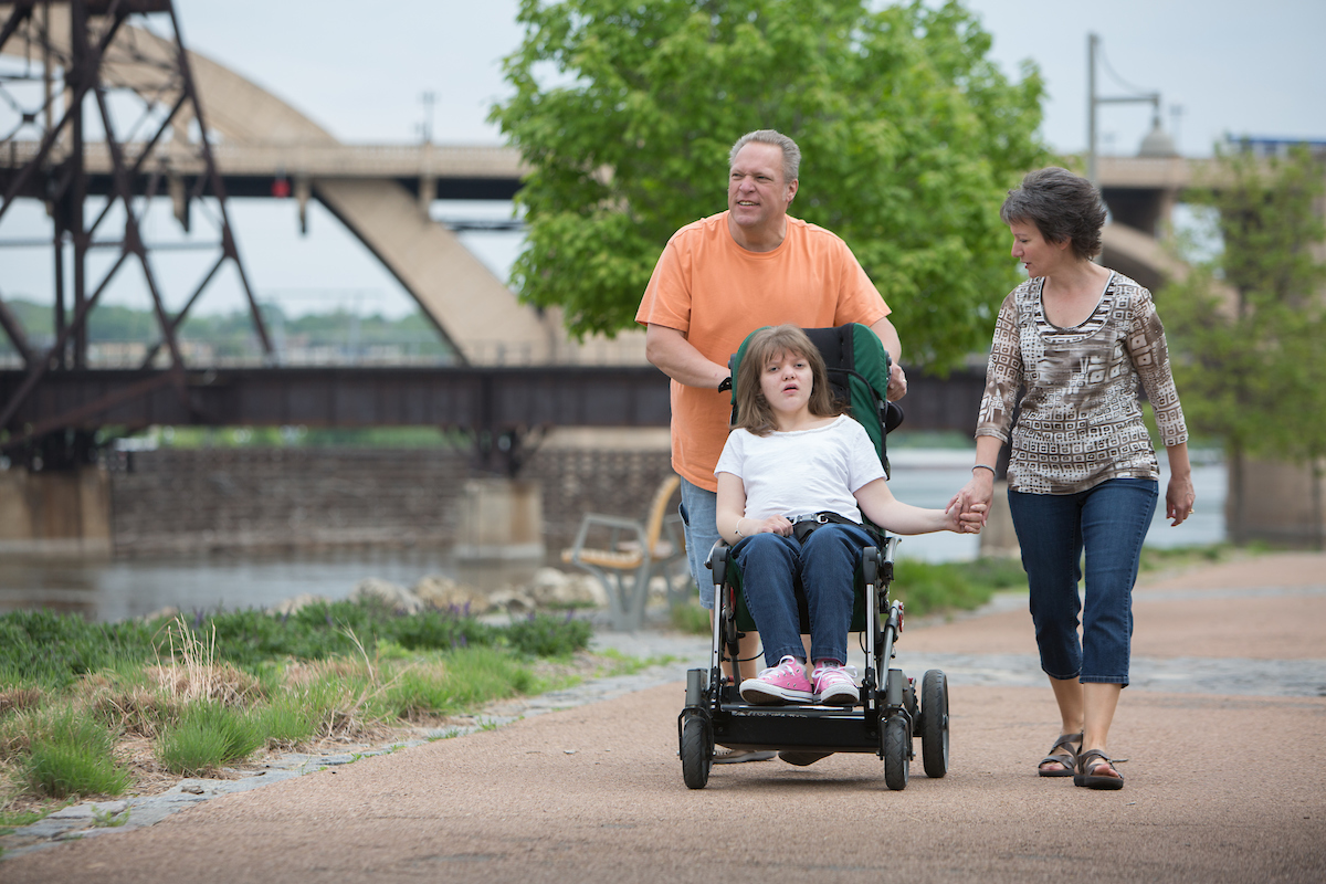 family of three walking on side walk pushing daughter in wheelchair 