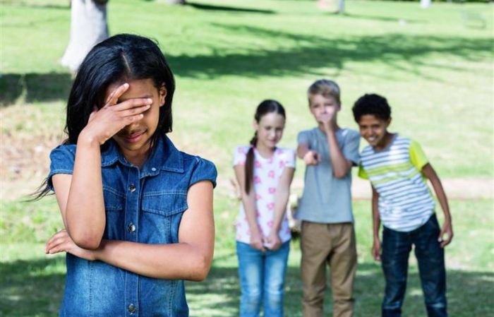A girl hides her face in her hand in the foreground as a group of three children in the background point and laugh.