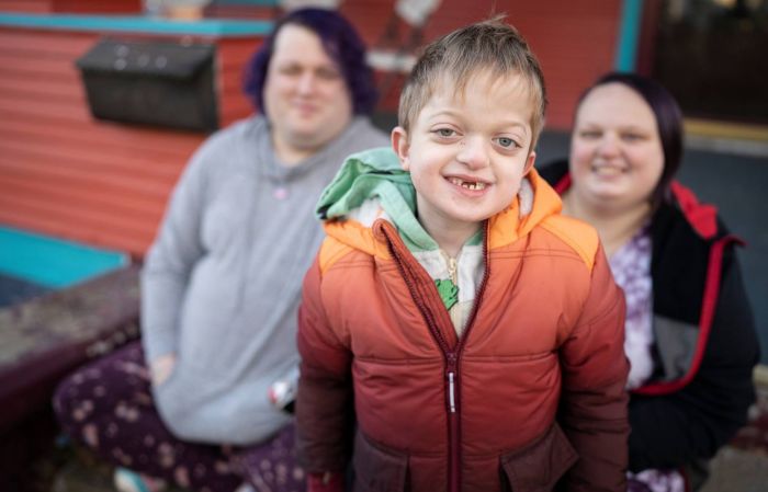 A gap toothed boy in an orange jacket smiles at the camera. Two adults are in the background.