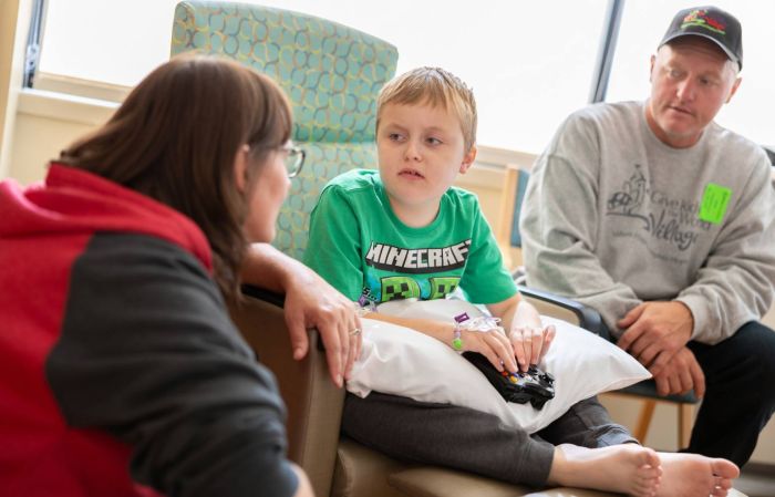 A boy in a green shirt sits on a chair with his mom to his right and his dad to his left in an infusion room at Gillette Children's.