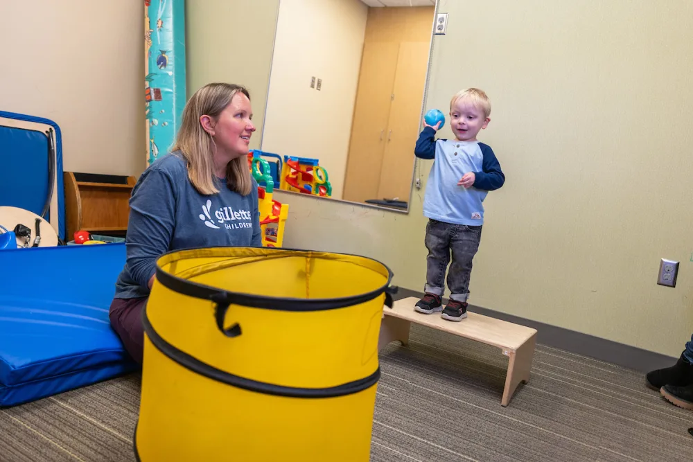 A young boy tries to throw a ball into a big yellow basket while his physical therapist encourages him.