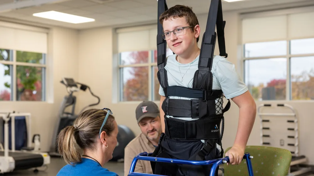 A teen boy is shown using a walker to steady himself as his body is strapped in to a physical therapy device to help him stand upright.