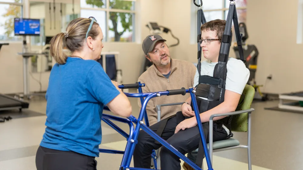 A teen boy is shown strapped into a ZeroG physical therapy device as he sits with his father and listens to a Gillette Children's rehabilitation therapist.