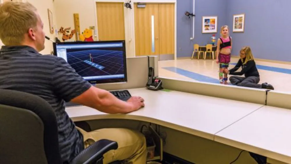 A young girl stands in the gait lab while a woman adjusts the senors on her legs and a male technician monitors her gait on a computer.