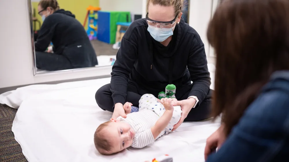 An infant lays on a white blanket with his head turned to the right while a woman dressed in black wearing a respiratory mask holds him still - Gillette Children's