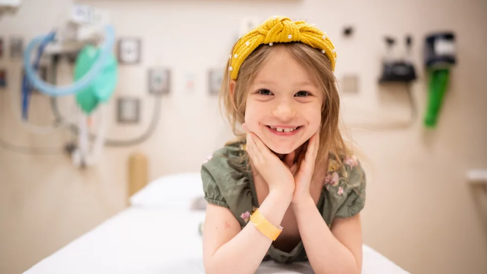 A five year old girl poses smiling while lying prone on a medical gurney with her chin resting on her hands.