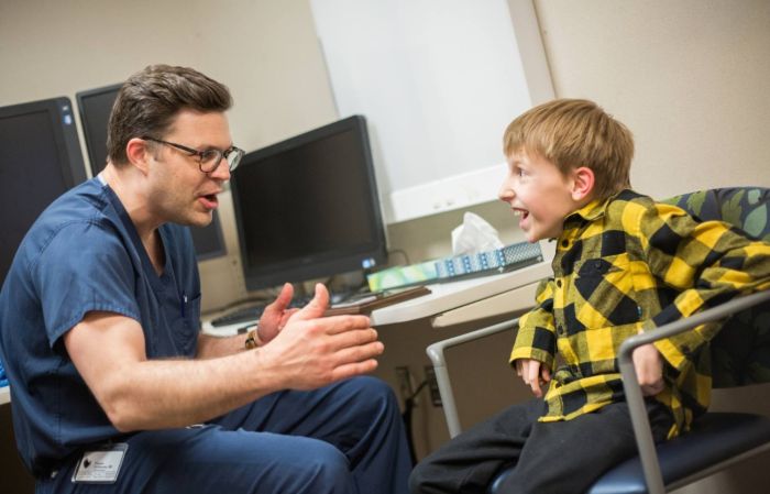 A doctor in scrubs talks to a smiling young boy who wears a checkered shirt in a medical exam room at Gillette Children's