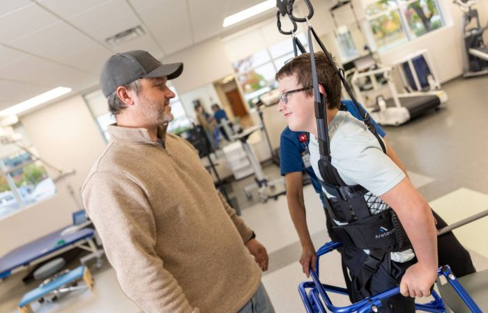 A boy using a walker and a ZeroG physical therapy system is shown from the side facing his father. The two are smiling at each other.