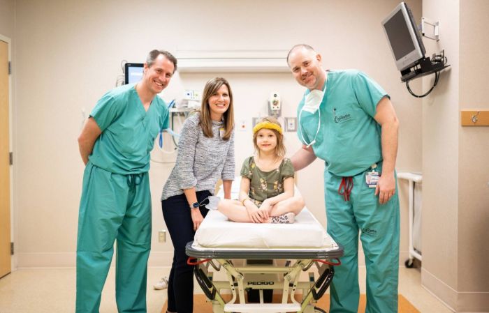 A five year old female patient poses smiling while seated on a medical gurney, surrounded by two men in surgical scrubs and her mother.