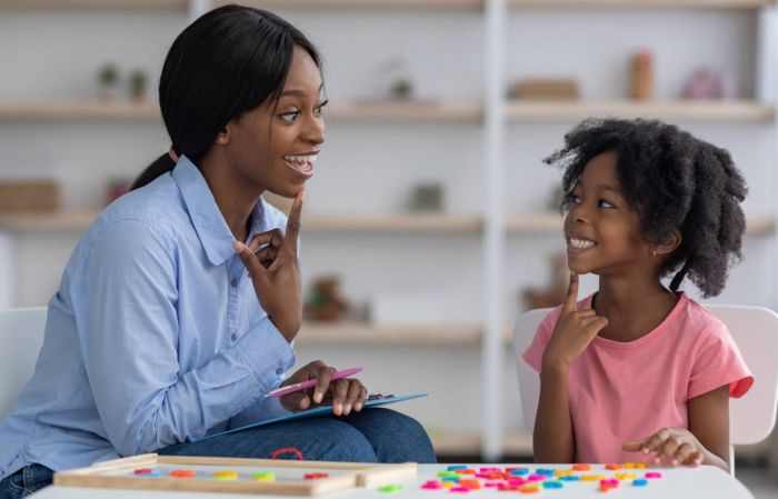 A young female in a pink shirt and an adult woman in a blue shirt point to their mouths as part of speech therapy at Gillette Children's.